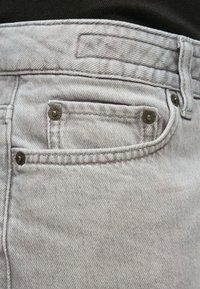 Lysegrå denimjeans har en frontlomme, synlige sømmer og metallknapper. Stoffet har en subtil tekstur og et lett falmet utseende.