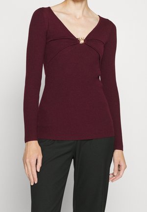 Kvinde iført en tætsiddende, ribstrikket bordeauxrød bluse med lange ærmer og en gulddetalje ved V-halsen, kombineret med sorte bukser, overkroppen synlig.