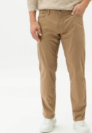 Mand står iført beige chinos, beige sweater og hvide sneakers, med den ene hånd i lommen og den anden afslappet ved siden.