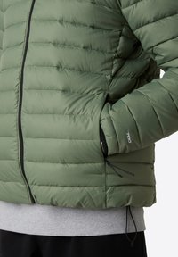 Giacca a vento in verde oliva, con chiusura a zip, cuciture orizzontali, tasche laterali e accessori neri. Superficie testurizzata.
