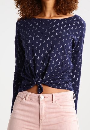 Woman wearing a navy blue long-sleeve top with white anchors, tied at the waist, and light pink jeans.