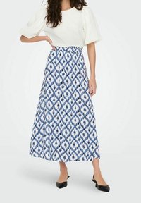 Blouse blanche texturée à manches bouffantes, associée à une jupe longue au sol bleue et blanche avec un motif en losanges et une taille cintrée.