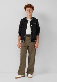 Zwarte varsityjacket met witte strepen, witte t-shirt, olijfgroene wijde broek en witte sneakers met beige zolen. Bevat een logopatch.