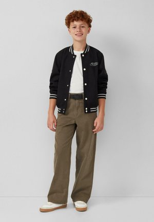 Veste varsity noire avec des rayures blanches, t-shirt blanc, pantalon large vert olive et baskets blanches avec des semelles beiges. Comprend un patch logo.