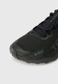Czarna sportowa buty z teksturowaną siateczką, wzmocnionym palcem i gumową podeszwą. Zawiera logo Gore-Tex i oznaczenie Lightstrike.