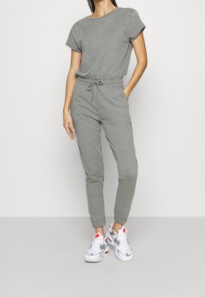 Kvinde stående iført en grå kortærmet jumpsuit med elastisk talje og hvide sneakers med røde detaljer.