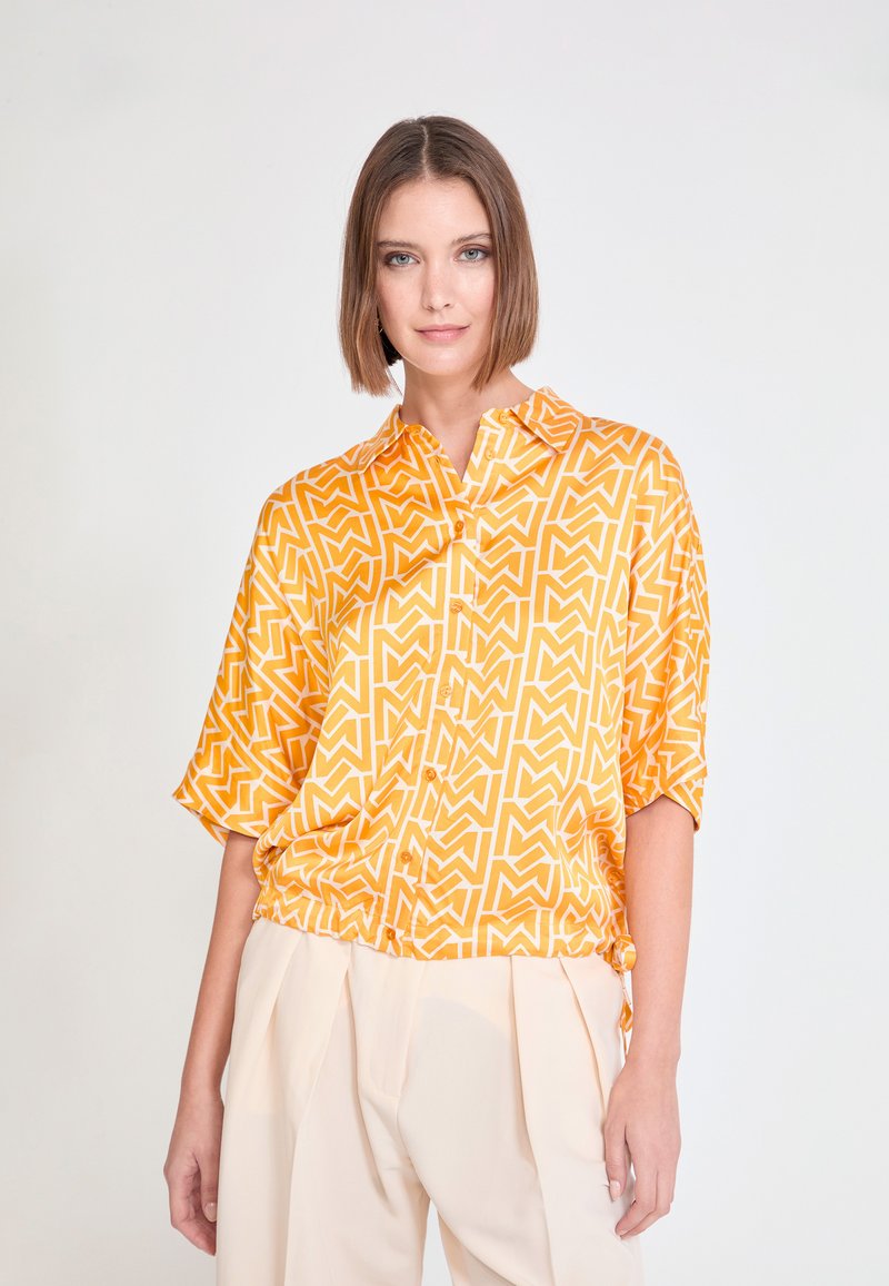 Chemise à manches courtes avec col, de couleur orange vif ornée d'un motif géométrique blanc. Tissu lisse avec une coupe décontractée, dotée de boutons sur le devant.