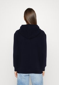 Navy hoodie with a drawstring, ribbed cuffs, and a relaxed fit. Soft fabric, features a large back pocket, worn over faded blue jeans.