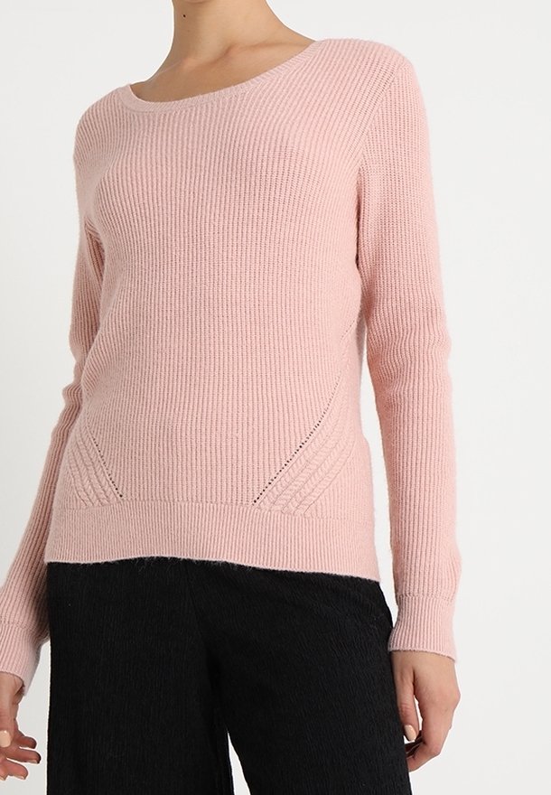 Femme portant un pull en maille côtelée rose clair à manches longues et un pantalon noir texturé sur fond blanc uni.