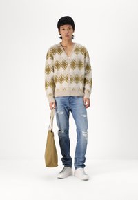 Cárdigan abotonado en crema con patrones de diamantes verdes; combinado con jeans claros rasgados y zapatillas blancas; llevando una bolsa de mano beige.