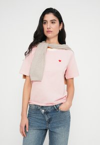 Ριγέ ροζ μπλουζάκι με κόκκινη καρδιά από ύφασμα, συνδυασμένο με ανοιχτό γκρι πουλόβερ που είναι περασμένο στους ώμους και μπλε τζιν.