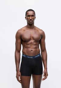 Črne bokserice z modrim pasom Calvin Klein. Tkanina deluje gladko, prileganje pa je tesno in oblikovano. Model stoji z sproščenimi rokami.