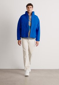 Uomo che cammina indossando una giacca blu, un maglione beige, pantaloni bianchi e sneakers bianche su uno sfondo bianco uniforme.
