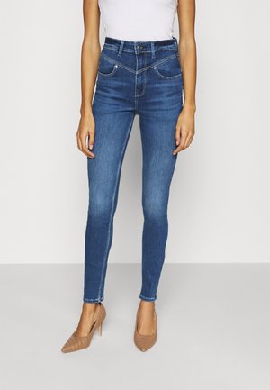 Femme portant un jean skinny bleu foncé taille haute avec des détails de coutures diagonales uniques et des escarpins beiges à bouts pointus, debout sur un fond uni.