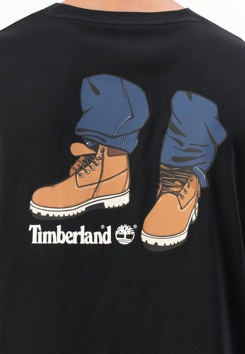 T-shirt nera con una grafica di stivali da lavoro marroni e pantaloni blu arrotolati. Logo Timberland in bianco sotto il design.