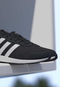 Sneaker Adidas bianca e nera con tre strisce bianche, lacci neri e suola bianca appoggiata su una superficie bianca.
