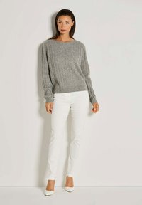 Frau steht vor einer weißen Wand und trägt einen grauen Kabelstrickpullover, weiße Hose und weiße spitze High Heels. Sie schaut leicht nach rechts.