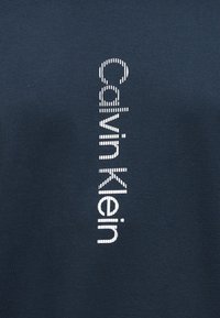 Σκούρο ύφασμα με κάθετο λευκό κείμενο "Calvin Klein" αποτελούμενο από σύντομες οριζόντιες γραμμές.