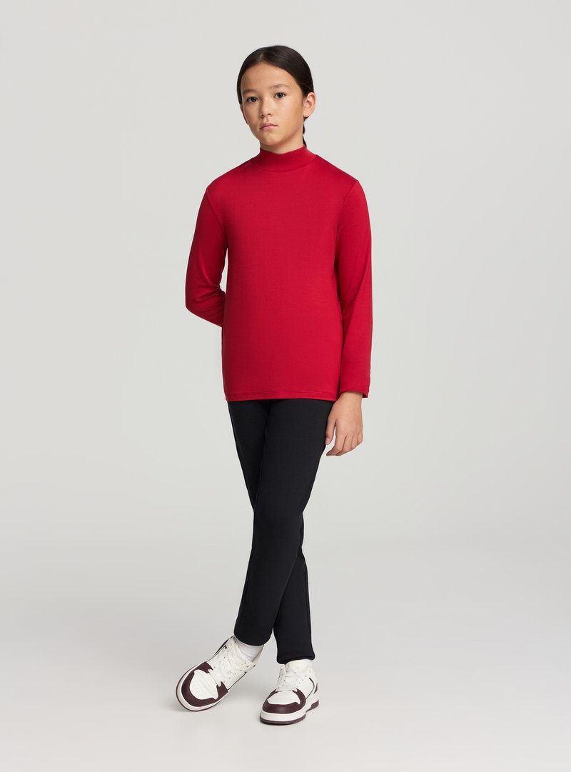 Bambino con capelli scuri, indossa una maglia rossa a maniche lunghe e pantaloni neri, in piedi con le braccia rilassate, con scarpe da ginnastica bianche e marroni su uno sfondo semplice.