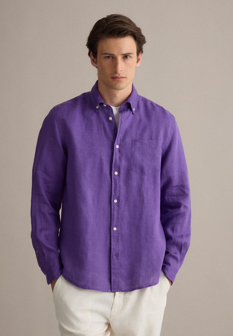 Joven con camisa morada de manga larga abotonada sobre una camiseta blanca y pantalones de color claro, de pie con las manos en los bolsillos.