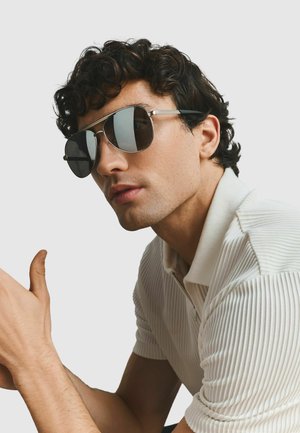 Mann mit dunklem lockigem Haar, der reflektierende Aviator-Sonnenbrillen und ein weißes, geripptes Poloshirt mit kurzen Ärmeln vor einem schlichten Hintergrund trägt.