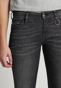 Vaqueros de mezclilla negra con un ajuste slim, diseño de cinco bolsillos y un acento de anillo dorado en la parte frontal. La tela tiene una textura suave con un ligero desvanecimiento.