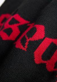 Sciarpa in tessuto a maglia nera con testo grafico rosso audace. La textura è liscia; il design presenta lettere pixelate con colori ad alto contrasto.