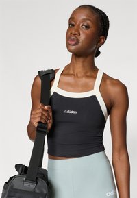 Juoda sportinė trumpa palaidinė su kreminėmis petnešomis, su gestuotas "adidas" logotipas. Modelis laiko juodą drobės krepšį, vilkėdamas šviesiai žalias pėdkelnes.