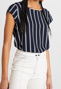 Blusa a righe blu navy con maniche corte, presenta strisce verticali bianche, design svasato, abbinata a pantaloni bianchi a vita alta.