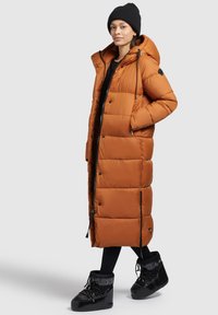 Lang, pufferjakke i brændt orange med hætte, trykknapper og quiltet design. Kombineret med sorte vandtætte støvler og en sort strikket hue.