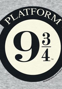 Schwarz-creamfarbiger runder Logo mit "PLATFORM 9¾", das eine große, stilisierte Zahl 9 und einen kleinen Bruch von drei Vierteln zeigt, vor einem grauen Stoffhintergrund.