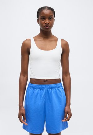 Γυναίκα με πλεγμένα μαλλιά που φοράει λευκό μπουστάκι χωρίς μανίκια και άνετο μπλε σορτς, στέκεται μπροστά σε απλό φωτεινό φόντο.