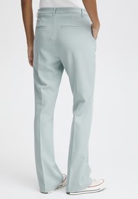 Persona vestida con pantalones de corte ligero en color azul claro, con bolsillos traseros de ojal y zapatillas blancas, de pie con una mano en el bolsillo.