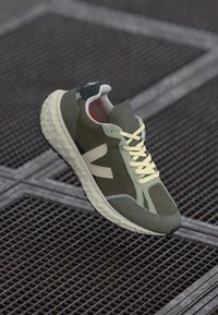 Zaļās sporta kurpes ar sieta paneļiem, teksturētām akcentiem, gaiši dzeltenām šņorēm un izteiktu zoli; uz sāniem ir redzams zīmola logo.