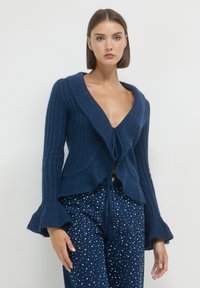 Femme portant un cardigan en tricot côtelé bleu foncé avec des manches évasées et un jean bleu foncé décoré de pois blancs sur un fond uni.