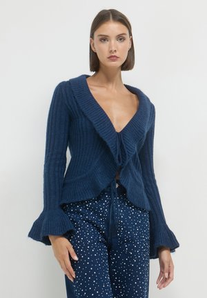 Femme portant un cardigan en tricot côtelé bleu foncé avec des manches évasées et un jean bleu foncé décoré de pois blancs sur un fond uni.