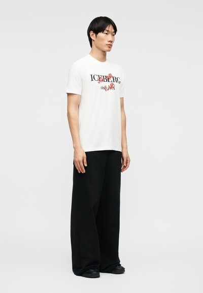 Νεαρός άνδρας με λευκό γραφικό μπλουζάκι με μαύρο και κόκκινο κείμενο και φαρδιά μαύρη παντελόνα, στέκεται πλευρικά μπροστά σε απλό φόντο.