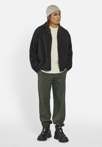 Black button-up jacket over a light beige t-shirt, paired with dark green striped pants and black boots. The model wears a beige knit beanie.