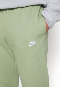 Persona vistiendo pantalones deportivos Nike verde claro con logo blanco en el muslo, mano en el bolsillo derecho y chaqueta de forro polar blanca.