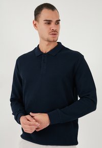 Polo cu mânecă lungă de culoare bleumarin, cu textură ribbed, guler, plachetă cu trei nasturi și manșete ajustate. Design simplu, fără modele.
