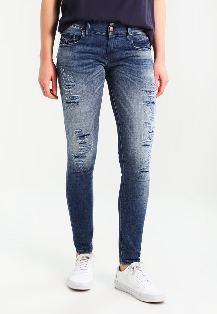 Personne portant un jean skinny bleu délavé et des baskets blanches, debout avec les mains détendues le long du corps sur un fond blanc.