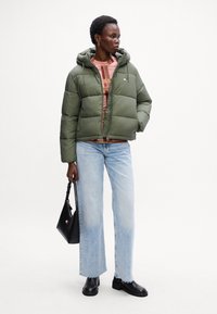 Grønn puffy jakke med hette, teksturert stoff, rutete topp, lyseblå jeans med slitte kanter, svarte sko og en liten svart veske.