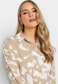 Blusa beige e bianca con motivo, colletto, chiusura con bottoni e una singola tasca sul petto; realizzata in tessuto morbido e fluente.