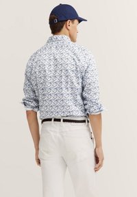Weißes Hemd mit blauem Blumenmuster, lange Ärmel, die an den Manschetten hochgerollt sind, und ein klassischer Kragen. Getragen mit weißen Hosen und einer blauen Mütze.