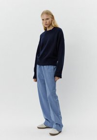 Mornarsko pleteno jopico s okroglim ovratnikom in spuščenimi rameni, v kombinaciji s svetlo modrimi karirastimi širokimi hlačami. Bele superge dopolnjujejo videz.