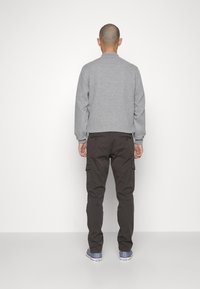 Giacca bomber trapuntata grigia con polsini a costine, abbinata a pantaloni cargo marrone scuro con tasche laterali. Scarpe casual completano il look.