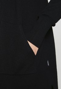 Maglione nero in maglia con texture a coste, tasca frontale e etichetta visibile. I polsini sono aderenti e il design è leggermente oversize.