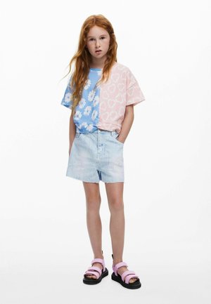 Camiseta de algodón azul con margaritas blancas en un lado y rosa con corazones blancos en el otro, combinada con pantalones cortos de mezclilla claros y sandalias rosas.