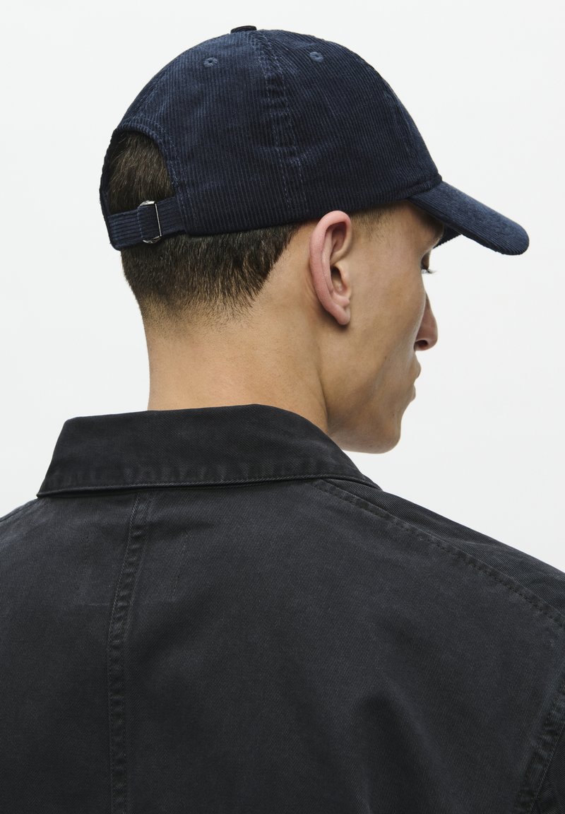 Navy corduroy cap with a curved brim and adjustable strap at the back, worn with a black collared jacket. Texture visible on the cap.