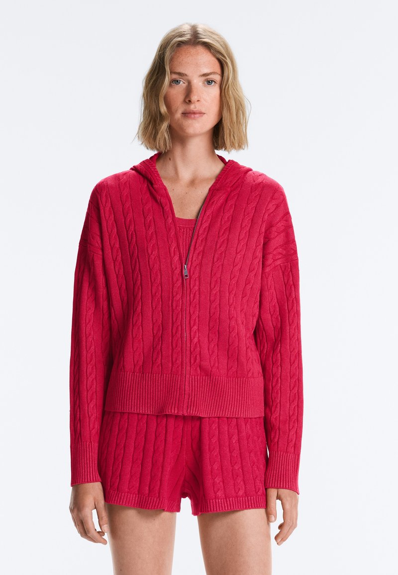 Femme portant un sweat à capuche rouge en maille torsadée avec zip et un short assorti, debout sur un fond gris clair.